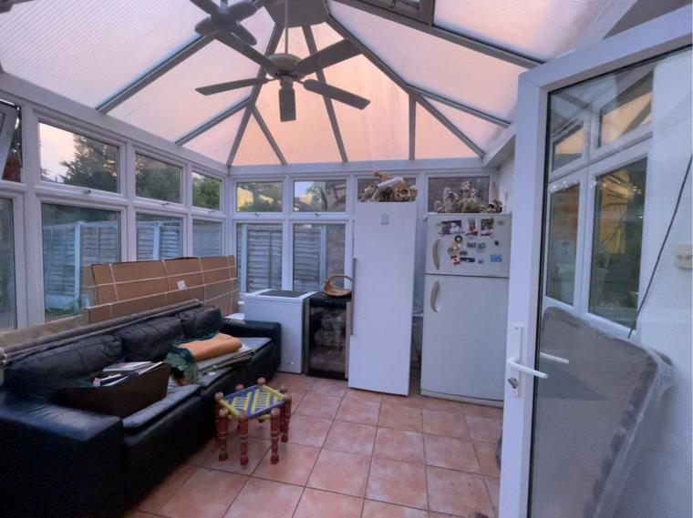 A dated uPVC conservatory with a pitched polycarbonate roof, tiled floor and an old sofa, being used for storage