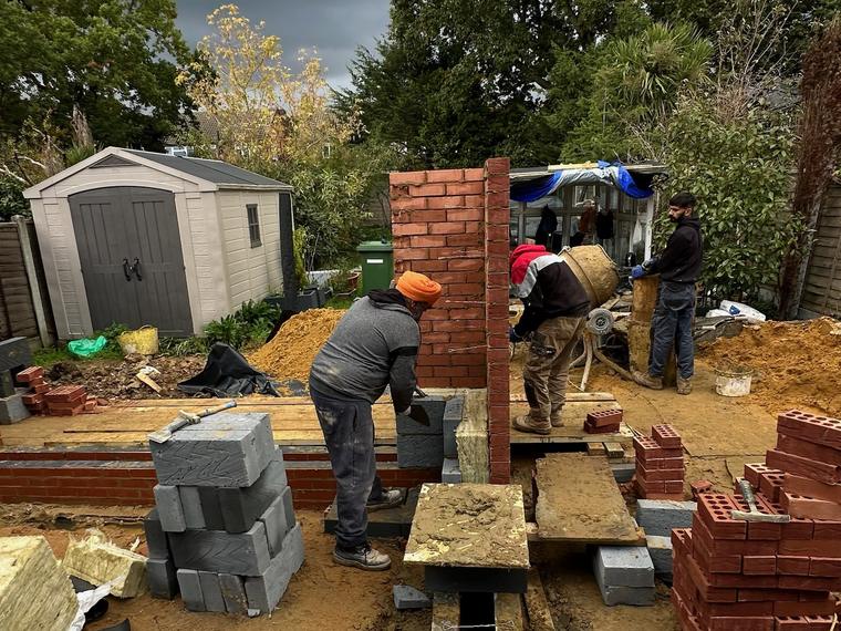 Groundworks in progress: trench foundations, a bricklayer working on a new pier, blockwork stacked and a cement mixer running under a dark sky
