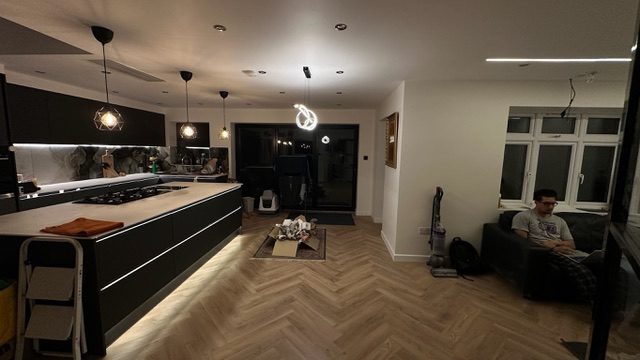 The finished open-plan kitchen: matte black handleless units, a large island with integrated hob, stone worktop, herringbone floor, pendant and recessed lighting