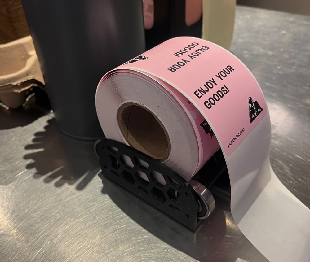 A 3D-printed black roller mount with a hexagonal lattice frame, holding a pink label roll on a bearing so the labels feed out flat across a steel counter